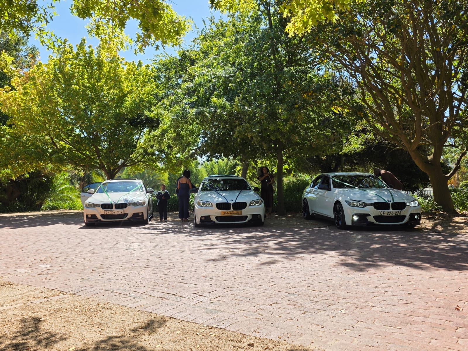 Ribboned white BMW wedding cars parked under trees at a garden venue